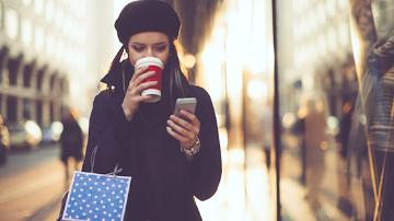 Female adult reading her mobile phone and drinking a coffee whilst walking and holding a shopping bag.