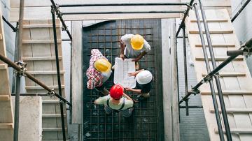 Vier Erwachsene auf einer Baustelle mit Schutzhelmen und Bauplänen, fotografiert von oben.