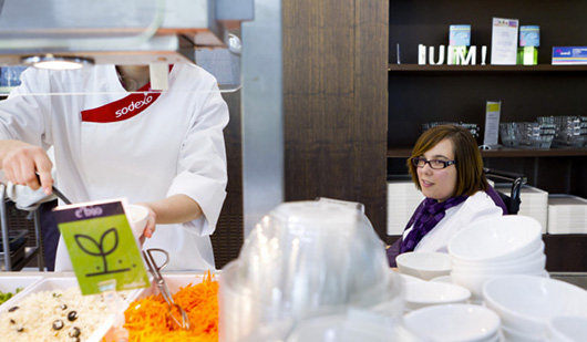Un employé SODEXO aide une personne en situation de handicap à se servir à un buffet de restaurant