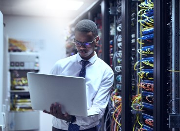 A professional man in a tie and glasses holds a laptop in a server room