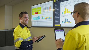Two facilities management employees talking, holding a tablet with screens showing data in the background