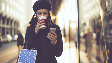Female adult reading her mobile phone and drinking a coffee whilst walking and holding a shopping bag